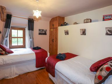 A bedroom with two single beds with red throws and pillows a wooden closet and a window with curtains at Tyddyn Morthwyl in Criccieth
