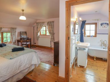 A bedroom with a bed, dressing table, and windows next to a bathroom with a bathtub and sink at Tyddyn Morthwyl in Criccieth
