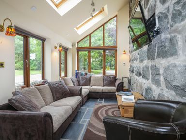 A living room with large windows two sofas an armchair a wooden coffee table a stone wall and a wall-mounted television at Tyddyn Morthwyl in Criccieth