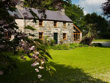 A stone house with a green door and several windows surrounded by a green lawn and trees at Tyddyn Morthwyl in Criccieth