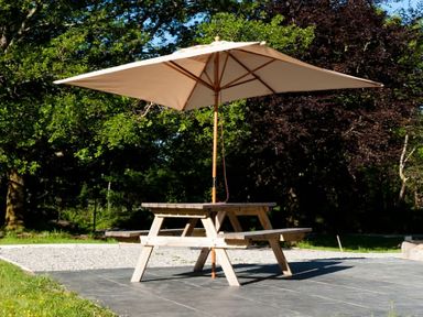 A wooden picnic table with a large beige umbrella on a patio surrounded by trees at Tyddyn Morthwyl in Criccieth