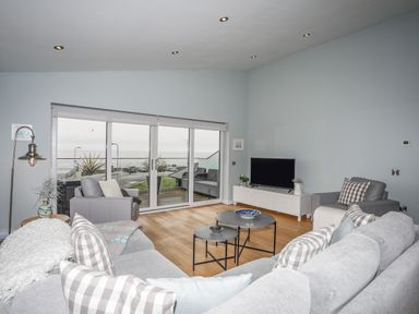 A living room with grey couches and checkered pillows and a TV on a stand near large glass sliding doors at Ty Glyn in Criccieth