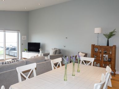A dining table with white chairs and vases with flowers in a living room with a sofa, armchairs, a TV, and a wooden cabinet at Ty Glyn in Criccieth