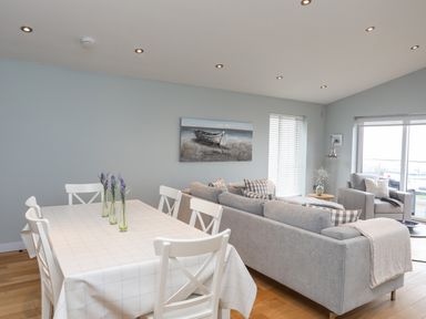 A dining table with six chairs and vases with flowers next to a living area with two sofas and armchairs at Ty Glyn in Criccieth