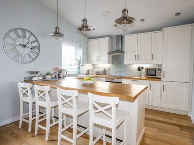 A kitchen with white cabinets a wooden countertop island with four white chairs and a large wall clock at Ty Glyn in Criccieth