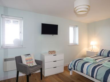 A bedroom with a bed striped bedding a bedside table with a lamp a white chest of drawers a wall-mounted television and a grey chair with a cushion at Ty Glyn in Criccieth