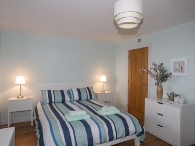 A bedroom with a double bed with striped bedding and two bedside tables with lamps and a dresser with a vase and cups at Ty Glyn in Criccieth