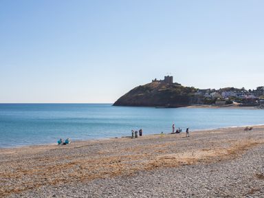 A pebble beach with people near the water and a castle on a hill in the background at Ty Glyn in Criccieth