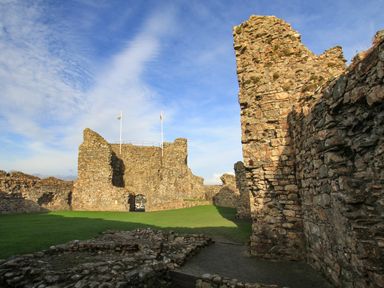 Stone ruins of a castle with two flagpoles on a grassy area under a blue sky at Ty Glyn in Criccieth
