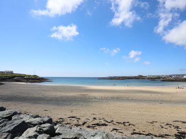 A sandy beach with rocks in the foreground people in the water and houses on the coast at Ty Llwyd in Trearddur Bay