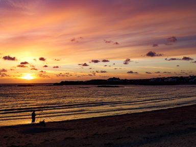 A person fishing on a beach at sunset with waves and houses in the distance at Ty Llwyd in Trearddur Bay