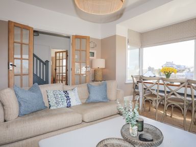 A living room with a beige sofa and blue pillows next to a dining table with chairs and yellow flowers at Ty Llwyd in Trearddur Bay