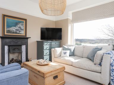 A living room with a beige sofa blue armchair wooden chest coffee table and a fireplace at Ty Llwyd in Trearddur Bay