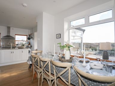 A dining table with chairs set with plates glasses candles and flowers near a kitchen with white cabinets and a large window at Ty Llwyd in Trearddur Bay