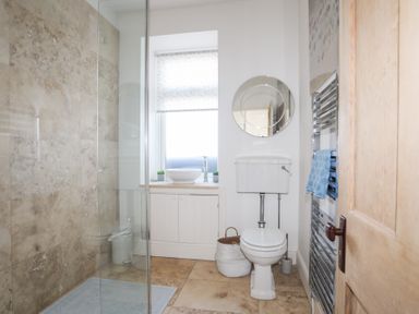 A bathroom with a glass shower enclosure a toilet a round mirror a towel rack and a sink by the window at Ty Llwyd in Trearddur Bay
