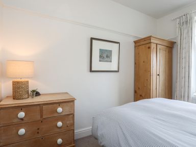 A bedroom with a wooden chest of drawers with a lamp on top a framed picture on the wall a wooden wardrobe and a bed with striped bedding at Ty Llwyd in Trearddur Bay