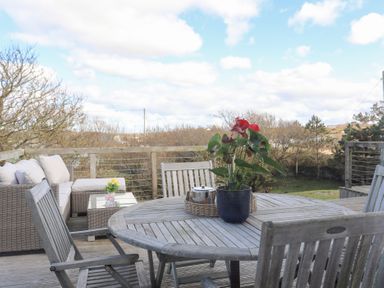 An outdoor wooden deck with a round wooden table and chairs a potted plant and a small tray with a teapot a wicker sofa with cushions and a glass coffee table with flowers in a vase at Ty Llwyd in Trearddur Bay