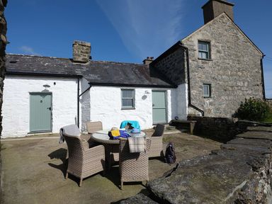 An outdoor patio area with a round glass table and six wicker chairs in front of a stone and whitewashed house at Ty Mawr Porthmadog in Porthmadog