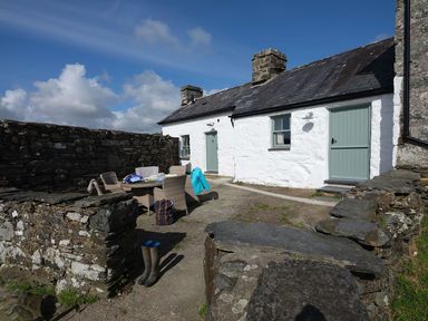 An outdoor patio area with a round table and four wicker chairs against a white stone cottage with two doors and two windows at Ty Mawr Porthmadog in Porthmadog