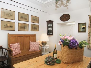 A room with a wooden bench and cushions a wooden table with flowers and a bottle six framed pictures on the wall a corner cabinet with dishes and a lamp on a small table at Ty Mawr Porthmadog in Porthmadog