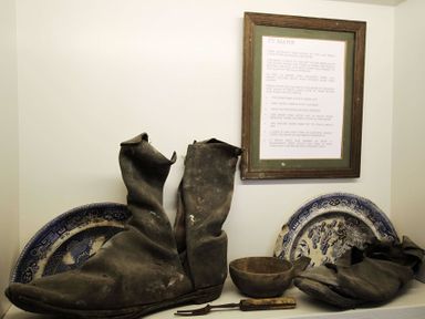 A display of old boots and vintage plates with a framed informational sign on a wall at Ty Mawr Porthmadog in Porthmadog