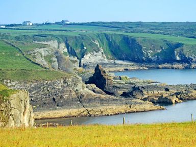 A coastline with rocky cliffs and grassy fields with small buildings on hills in the background at Ty Nant in South Stack