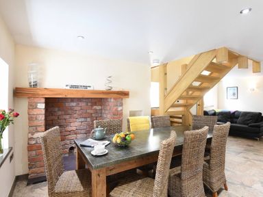 A dining area with a wooden table and wicker chairs next to a brick fireplace and wooden staircase at Ty Nant in South Stack