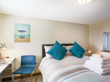 A bedroom with a bed with blue pillows a stack of white towels a blue chair a desk with lamps and a beach-themed picture on the wall at Ty Nant in South Stack