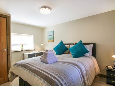 A bedroom with a bed with striped bedding and teal pillows folded towels on the bed a wooden door a window a desk with a lamp and decor at Ty Nant in South Stack