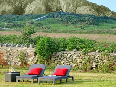 Two outdoor lounge chairs with red pillows on grass near a stone wall with shrubs and a wooden bench at Ty Nant in South Stack