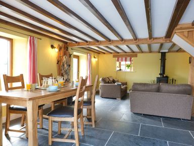 A dining area with a wooden table set for breakfast and a living area with sofas and a wood stove at Tyn Llwyn Cornel in Beddgelert