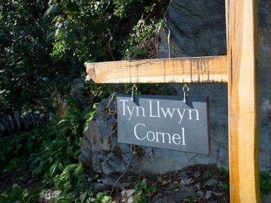 A wooden post with a hanging sign reading Tyn Llwyn Cornel surrounded by rocks and plants at Tyn Llwyn Cornel in Beddgelert