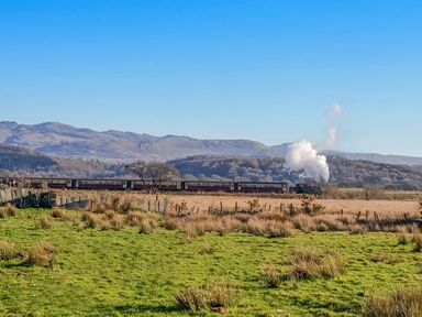 A steam train traveling across a field with mountains in the background at Tyn Llwyn Cornel in Beddgelert