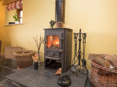 A wood burning stove with fire tools and baskets of logs near a window at Tyn Llwyn Cornel in Beddgelert
