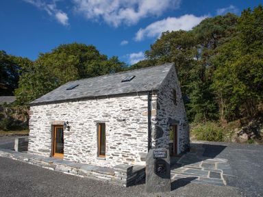 A stone cottage surrounded by trees with a paved pathway and a sign reading Tyn Llwyn Cornel at Tyn Llwyn Cornel in Beddgelert