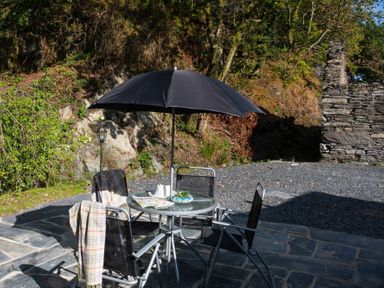 An outdoor patio with a round glass table and four chairs under a black umbrella at Tyn Llwyn Cornel in Beddgelert