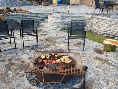 An outdoor stone patio with a fire pit cooking meat and chicken surrounded by black mesh chairs and stacked firewood at Tyn Llwyn Cornel in Beddgelert