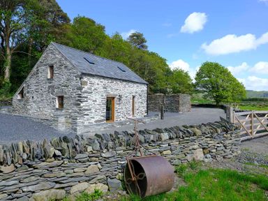 A stone house with a slate roof next to a stone wall and a metal roller by a wooden gate in a rural area at Tyn Llwyn Cornel in Beddgelert
