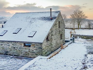 A stone house with a snow-covered roof and ground with trees and fields in the background at Tyn Llwyn Cornel in Beddgelert