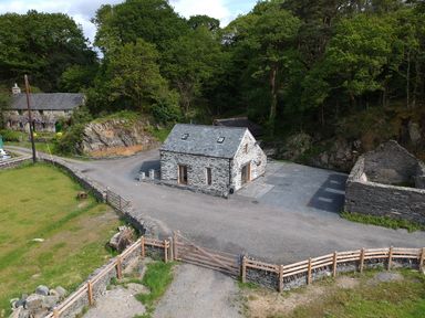 A stone cottage with a slate roof surrounded by trees and gravel driveway at Tyn Llwyn Cornel in Beddgelert