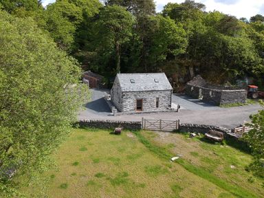 A stone cottage with a slate roof near trees and a grassy area at Tyn Llwyn Cornel in Beddgelert