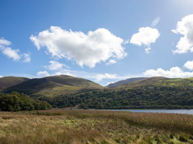 A landscape with grassy fields trees a lake and hills under a blue sky at 17 Victoria Terrace - Perthi in Nantlle