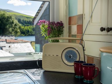 A kitchen countertop with a retro radio two mugs flowers and a window showing rooftops and greenery at 17 Victoria Terrace - Perthi in Nantlle