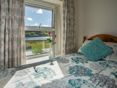 A bedroom with a wooden bed a blue cushion floral curtains and two vases with pink flowers by the window at 17 Victoria Terrace - Perthi in Nantlle