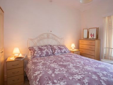 A bedroom with a bed with purple floral bedding two bedside tables with lamps a wooden chest of drawers and a chair at 17 Victoria Terrace - Perthi in Nantlle