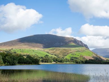 A lake with trees and grassy hills with a mountain in the background under a blue sky with clouds at 17 Victoria Terrace - Perthi in Nantlle