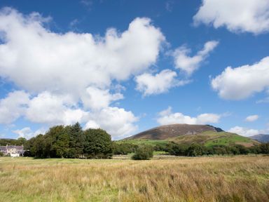 A grassy field with trees and a hill under a blue sky with clouds at 17 Victoria Terrace Perthi in Nantlle