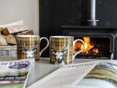 Two mugs with deer illustrations on a table near a burning wood stove with stacked firewood and magazines at Mulberry Cottage in Pentraeth