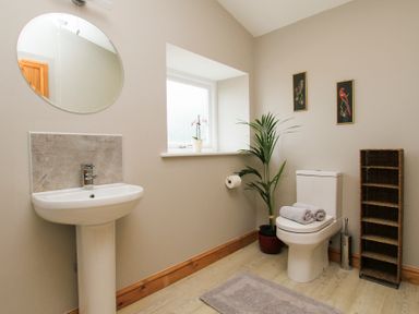 A bathroom with a pedestal sink a round mirror a toilet with towels a plant a window and a shelving unit at Mulberry Cottage Pentraeth