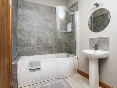 A bathroom with a bathtub shower combination a sink with a round mirror and grey tiled walls at Mulberry Cottage in Pentraeth